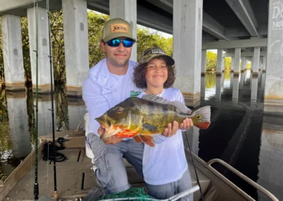 A person and a child proudly hold a large fish in a boat beneath a concrete bridge surrounded by lush greenery