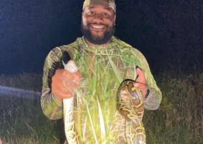 A person in camouflage clothing holds a large python in one hand and a smaller snake in the other, outdoors at night.